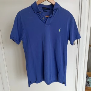 Blå pikétröja från Polo Ralph Lauren - Klassisk blå pikétröja från Polo Ralph Lauren med gul broderad logga på bröstet. Tröjan har korta ärmar, krage och knappar vid halsen. Perfekt för en stilren look.