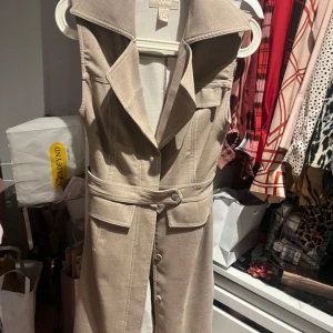 Väst klänning  - Snygg beige ärmlös klänning med krage och knappar framtill. Klänningen har markerad midja med avtagbart bälte och fickor framtill. Perfekt för en stilren och modern look.