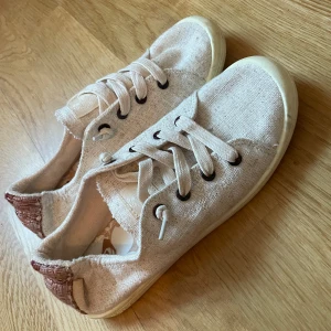 Beige sneakers med snörning och brun detalj - Snygga beige sneakers med breda snören och bruna detaljer vid hälen. Skorna har rund tå och platt sula, tillverkade i textilmaterial med dekorativ söm baktill. Perfekta för en avslappnad stil.