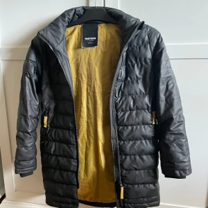 Svart pufferjacka från Tretorn - Svart pufferjacka från Tretorn med gul insida och huva. Jackan har dragkedja framtill och gula detaljer vid fickorna. Perfekt för kyliga dagar och har en modern, sportig look.