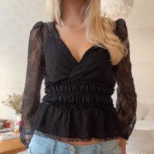 Svart spetsblus med volang - Säljer en svart blus i spets med långa ärmar och omlott-skärning. Blusen har volang nertill och smockad midja som ger en feminin siluett. Perfekt att styla med jeans eller kjol för en trendig look.