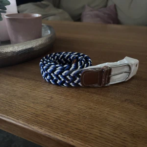 Flätat blåvitt bälte med bruna skinn-detaljer - Säljer ett snyggt flätat bälte i blått, vitt och svart med bruna skinn-detaljer vid spännet. Bältet har ett silverfärgat metallspänne och passar perfekt till jeans eller chinos.
