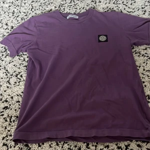 Lila t-shirt från Stone Island Junior - Säljer en lila t-shirt från Stone Island Junior med klassisk logotyp-patch på bröstet. T-shirten har rund hals och är tillverkad i mjuk bomull. Perfekt för dig som gillar streetwear och vill ha något stilrent men ändå unikt.