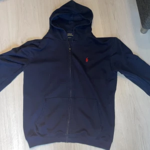Mörkblå hoodie med dragkedja från Polo Ralph Lauren - Säljer en mörkblå hoodie från Polo Ralph Lauren med dragkedja och huva. Tröjan har en röd broderad logga på bröstet och två fickor framtill. Perfekt för en avslappnad stil. Storleken är XL men sitter som L/M. Jag på bilden som har på mig den är 187 och väger 80 kilo.