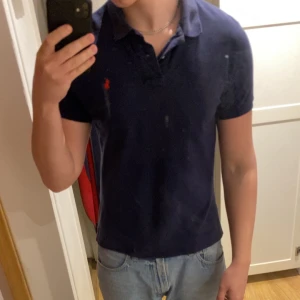 Mörkblå pikétröja från Ralph Lauren - Säljer en klassisk mörkblå pikétröja från Ralph Lauren med röd broderad logga på bröstet. Tröjan har krage, korta ärmar och knäppning vid halsen. Perfekt för en stilren och avslappnad look.