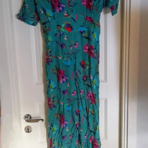 Turkos blågrön blommig byxdress från Dilvin - Säljer en turkos byxdress från Dilvin med färgglatt blommigt mönster. Den har korta ärmar och en avslappnad passform, perfekt för vår och sommar. Materialet är lätt och luftigt, vilket gör den skön att bära under varma dagar. Oanvänd. Bara upptvättad 