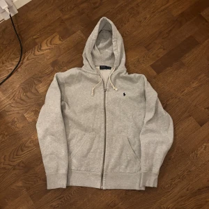 Grå hoodie med dragkedja från Polo Ralph Lauren - Säljer en klassisk grå hoodie från Polo Ralph Lauren i storlek M. Tröjan har dragkedja, huva med snörning och två fickor framtill. Liten broderad logga på bröstet. Perfekt för en avslappnad stil. Passar S. Det är brorsans och den blev för liten för honom så han gav den till mig. Jag vill inte ha den för det är inte riktigt min stil. Inga defekter😁Pris kan diskuteras 