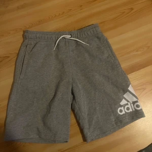 Grå shorts från Adidas med snörning - Grå mjuka shorts från Adidas med vit snörning i midjan och stort vitt Adidas-tryck på ena benet. Shortsen har sidofickor och en bakficka, perfekt för en avslappnad stil.