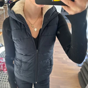 Svart dunväst med huva och foder - Svart dunväst med dragkedja framtill och mysigt ljust foder i kragen. Västen har en normal passform och en stor huva.