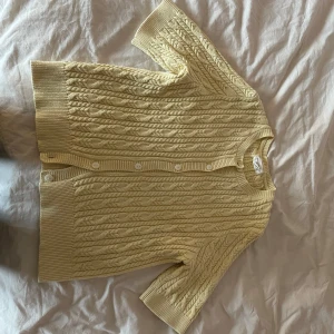 Gul kabelstickad kofta från Holly & Whyte - Säljer en ljusgul kabelstickad kofta från Holly & Whyte med korta ärmar och knappar framtill. Tröjan har rund halsringning och ribbade kanter. Perfekt för lager-på-lager och en snygg detalj till din outfit.