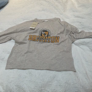 Grå croppad långärmad tröja från Mango - Säljer en ljusgrå croppad långärmad blus från Mango med tryck i gult och svart där det står 'BRIGHTON' och ett emblem framtill. Perfekt för en avslappnad och trendig look.