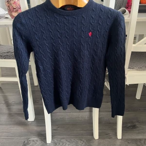 Mörkblå kabelstickad tröja från Morris - Säljer en stilren mörkblå kabelstickad tröja från Morris i 100% merinoull. Tröjan har en liten röd broderad detalj på bröstet och långa ärmar. Perfekt för kyligare dagar. Den är liten i storleken så den passar mer xs/s