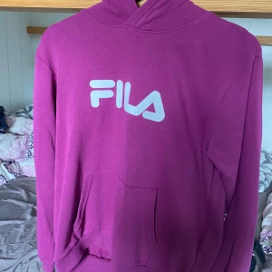 Lila hoodie från FILA - Säljer en lila hoodie från FILA med vit logga på bröstet. Tröjan har huva och en stor magficka framtill. Perfekt för dig som gillar sportiga och bekväma plagg.