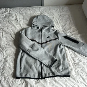 Nike tech fleece - Snygg grå ziphoodie från Nike med svart logga på bröstet och svarta detaljer. Jackan har huva, hel dragkedja framtill och en praktisk ficka med dragkedja på ärmen. Perfekt för en sportig och avslappnad stil. Pris kan diskuteras vid snabb affär 💕