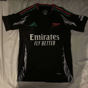 Arsenal bortatröja 24/24 fotbollströja  - Säljer en svart fotbollströja från Adidas med korta ärmar och snygga detaljer i vitt och rött. Tröjan har en modern design med klubbmärke och sponsorlogga på framsidan. Perfekt för fotbollsfans som vill ha en stilren look på planen.