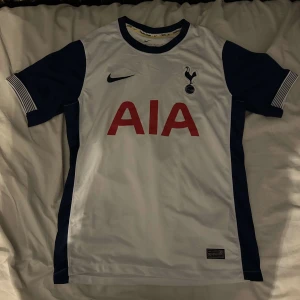 Tottenham hotspur hemmatröja 24/25 fotbollströja  - Säljer en vit fotbollströja från Nike med mörkblå detaljer och korta ärmar. Tröjan har en röd AIA-logga på framsidan och en klubbemblem på bröstet. Perfekt för fotbollsfans!