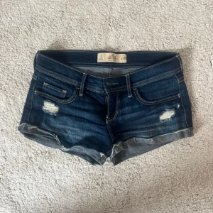 Jeansshorts från Hollister - Snygga mörkblå jeansshorts med slitna detaljer och uppvikta kanter. Perfekta för en avslappnad sommarlook. De har en klassisk femficksdesign och knappgylf.