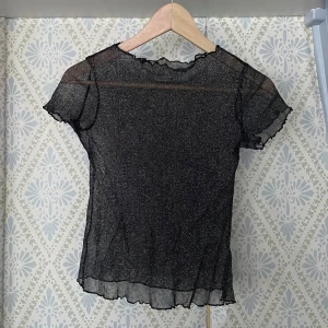 Transparent topp - Säljer en svart transparent glittrig topp med korta ärmar och volangdetaljer vid ärmslut och nederkant. 
