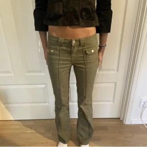 Olivgröna byxor med fickdetaljer - Snygga olivgröna byxor med låg midja och raka ben. Byxorna har framträdande sömmar och knappar vid fickorna som ger en cool look. Perfekta för en avslappnad stil.