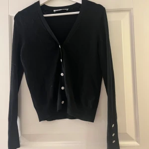 Svart kofta med knappar i silver  - Snygg svart kofta med v-ringning och dekorativa knappar framtill. Långärmad design med knappar även vid ärmsluten. Perfekt för en stilren look. Strl S. Köpt från Zara. Har knappt aldrig använt den.