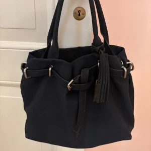 Svart axelväska med tofsdetalj - Snygg svart axelväska med stilren design och tofsdetalj. Väskan har ett knytband och metallringar som ger en modern touch. Perfekt för att bära dina dagliga nödvändigheter med stil.