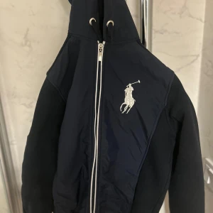 En OG marinblå/mörkblå Raulph Laurent cardigan. Full zip med luva. Perfekt flr en stilren look. Bra skick och finns inte längre att köpa. - Snygg mörkblå/marinblå cardigan från Ralph Lauren med dragkedja och broderad logga på bröstet. En OG marinblå/mörkblå Raulph Laurent cardigan. Full zip med luva. Perfekt för en stilren look. Bra skick och finns inte längre att köpa. Köpte den men satt lite litet på mig så säljer iväg den utan att jag har använt den.