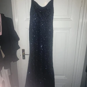 Mörkblå glittrig långklänning - Säljer en elegant mörkblå långklänning med glittrande detaljer. Klänningen har smala axelband och en figurnära passform som ger en stilren look. Perfekt för speciella tillfällen.