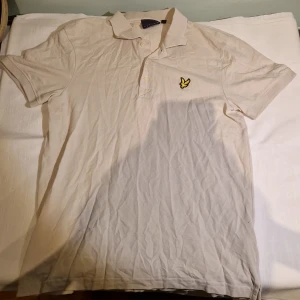 Vit pikétröja från Lyle & Scott - Säljer en klassisk vit pikétröja från Lyle & Scott med korta ärmar och en broderad gul logotyp på bröstet. Tröjan har en krage med knappar och är perfekt för en stilren look. Perfekt till vår och sommaren.