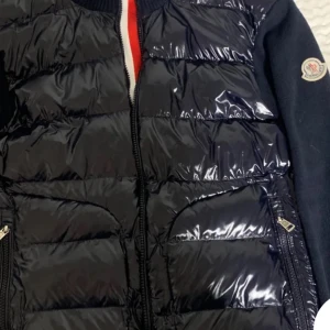 Svart/mörkblå cardigan från Moncler - Snygg svart pufferjacka från Moncler med glansigt material och dragkedja framtill. Jackan har långa ärmar och en diskret logga på ärmen. Perfekt för kyligare dagar.