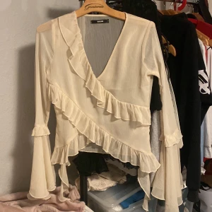 Beige omlottopp från BikBok - Säljer en elegant beige omlottopp från BikBok med volangdetaljer. Toppen har långa ärmar och en v-ringad design som ger en stilren look. Perfekt för att lyfta vilken outfit som helst! Har en liten fläck på en av volangerna men det syns knappast
