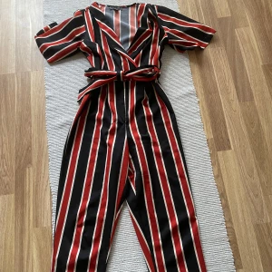 Randig jumpsuit med knytband - Snygg randig jumpsuit i svart, rött och vitt med korta ärmar och v-ringning. Den har ett knytband i midjan för en justerbar passform. Perfekt för en trendig look!