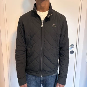 Quilted Jacka från GANT - Passar både XS och S. Snygg svart quiltad jacka från GANT med dragkedja och hög krage. Perfekt för kyligare dagar med sin stilrena design och diskreta logga på bröstet. Jackan har långa ärmar och en klassisk passform.