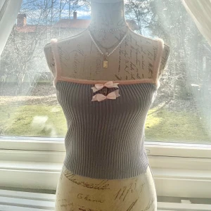 Grå ribbad linnetopp med rosa detaljer - Söt grå ribbad linnetopp med rosa kanter och en liten rosett framtill. Perfekt för en avslappnad stil med en touch av gullighet. Färgen är lite mellan grå-blå.