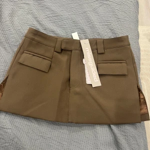 Brun kortkjol från Missguided - Snygg brun kortkjol från Missguided med stilrena fickdetaljer framtill. Kjolen har en klassisk design med bälteshällor och en rak passform. Perfekt för att skapa en chic look. Kjolen är i storlek S och helt oanvänd.