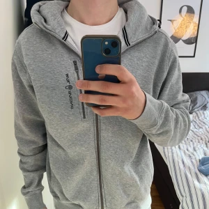Grå hoodie från Sail Racing - Säljer en stilren grå hoodie från Sail Racing med dragkedja framtill i storlek M. Nypris:1400kr