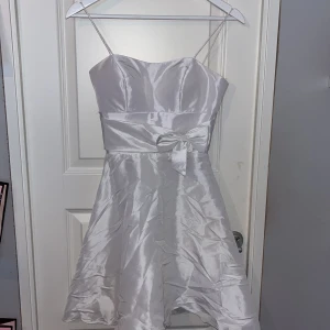 Vit axelbandslös klänning med rosett - Elegant vit klänning i satin med smala axelband och en söt rosett i midjan. Perfekt för speciella tillfällen med sin glänsande yta och stilrena design.