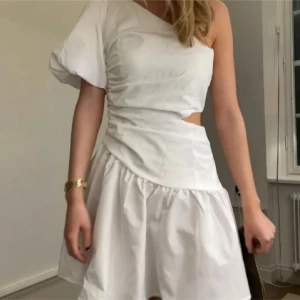 Vit studentklänning  - Snygg vit oneshoulder klänning med en unik design och cut-out detalj i midjan. Den har en kort och fladdrig kjol perfekt för sommarens alla tillfällen tex. Studenten. Storlek S