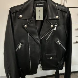 Svart skinnjacka från Balenciaga - Snygg svart skinnjacka från Balenciaga med coola dragkedjedetaljer och tryck på ryggen. Jackan har en klassisk bikerstil med knappar och flera fickor. Perfekt för att ge din outfit en edgy touch.