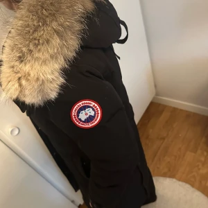 Svart dunjacka med päls från Canada Goose - Säljer en svart dunjacka från Canada Goose med en lyxig pälsdetalj på huvan. Jackan har en lång passform och är utrustad med både dragkedja och knappar för extra skydd mot kylan. Perfekt för kalla vinterdagar. ( knappen längst ner går inte att stänga ) ! 
