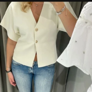 Vit kortärmad blus med knappar - Snygg vit blus med korta ärmar och två dekorativa knappar framtill. Perfekt för en stilren look.