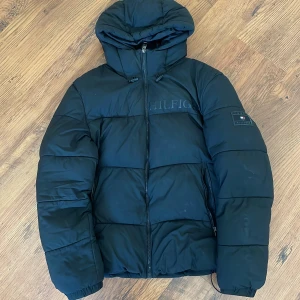 Tommy Hilfiger jacka - Säljer en svart pufferjacka från Tommy Hilfiger med huva och dragkedja. Jackan har märkets logga diskret på bröstet och en patch på ärmen. Perfekt för kyliga dagar.