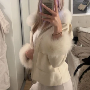 Vit jacka med pälsdetaljer - Säljer en stilren vit jacka med fluffiga pälsdetaljer på krage och ärmslut. Jackan har en dragkedja framtill och en elegant passform som ger en lyxig känsla. Perfekt för kyligare dagar när du vill hålla stilen.