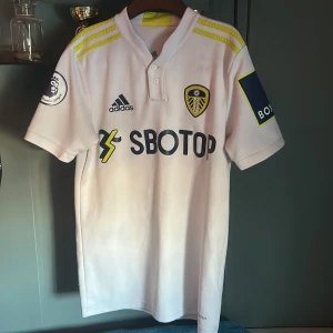 Vit Leeds United Raphina fotbollströja från Adidas - Säljer en vit Leeds United raphina fotbollströja från Adidas med korta ärmar. Tröjan har gula detaljer och sponsorloggor. Raphinha och nummer 10 är tryckt på baksidan. Perfekt för fans av laget!