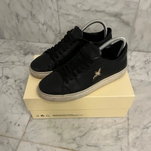 Axel Arigato skor - Snygga svarta sneakers från Axel Arigato med en broderad fågel på sidan. Skorna har en klassisk design med snörning och en vit. Skick 9/10 förutom att hälen på skon är lite slapp men sparsamt använda, nypris runt 2500kr. Pris kan diskuteras vid snabb affär