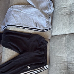 Adidas mjukisbyxor i grått och svart - Två par Adidas mjukisbyxor, ett i grått och ett i svart. Båda har de klassiska tre ränderna längs sidorna och elastisk midja för bekväm passform. Perfekta för en avslappnad stil. Svarta Storlek L och gråa storlek M