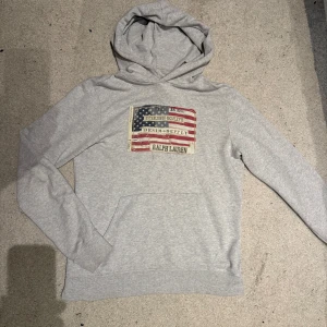 Grå hoodie från Ralph Lauren - Säljer en stilren grå hoodie från Ralph Lauren med ett tryck av den amerikanska flaggan och texten 'Denim & Supply'. Tröjan har en klassisk bekväm huva.