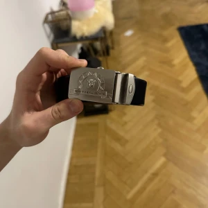 Svart bälte från Versace - Äkta vintage !!! Snyggt svart bälte från Versace med en iögonfallande silverfärgad spänne. Bältet är tillverkat i skinn och har ett klassiskt och stilrent utseende. Perfekt för att ge en lyxig touch till din outfit.