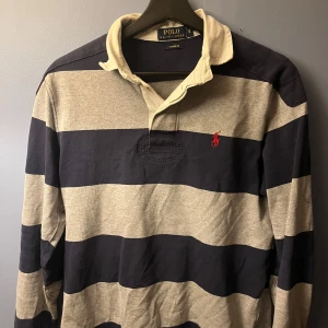 Långarmad piketröja från Ralph Lauren - Säljer en klassisk randig pikétröja från Ralph Lauren i marinblått och beige. Tröjan har långa ärmar och en krage i kontrasterande färg. Den ikoniska röda loggan är broderad på bröstet. Perfekt för en stilren look!