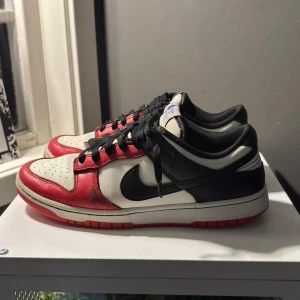 Nike Dunk i rött, svart och vitt - Snygga Nike Dunk sneakers i en klassisk färgkombination av rött, svart och vitt. Skorna har en låg profil med svart snörning och en vit sula med röd undersida. Perfekta för en stilren look.