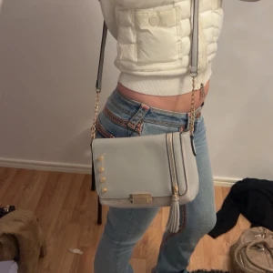 Ljusgrå axelväska med gulddetaljer från River Island - Säljer en stilren ljusgrå axelväska från River Island med guldfärgade nitar och kedja. Väskan har dragkedja, dekorativ tofs och en metallplatta framtill. Perfekt storlek för det viktigaste och passar till många outfits.